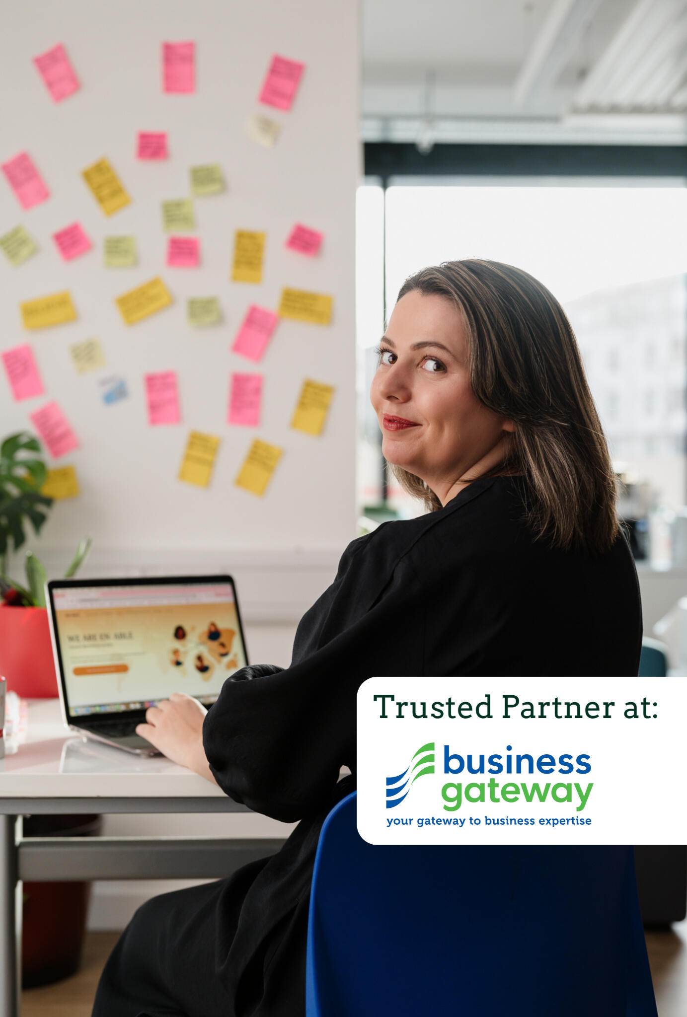 Kitti Majoran is sitting in an office at a desk with a laptop, looking back at the camera. In the bottom right corner of the photo, the writing 'Trusted Partner at:' can be seen with a logo of the Business Gateway and their slogan: 'Your Gateway to Busines