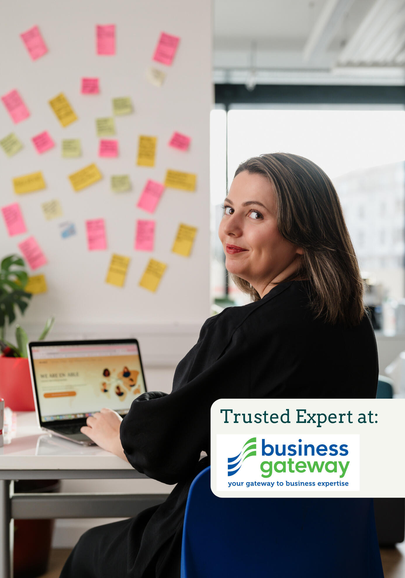 Kitti Majoran is sitting in an office at a desk with a laptop, looking back at the camera. In the bottom right corner of the photo, the writing 'Trusted Partner at:' can be seen with a logo of the Business Gateway and their slogan: 'Your Gateway to Busines