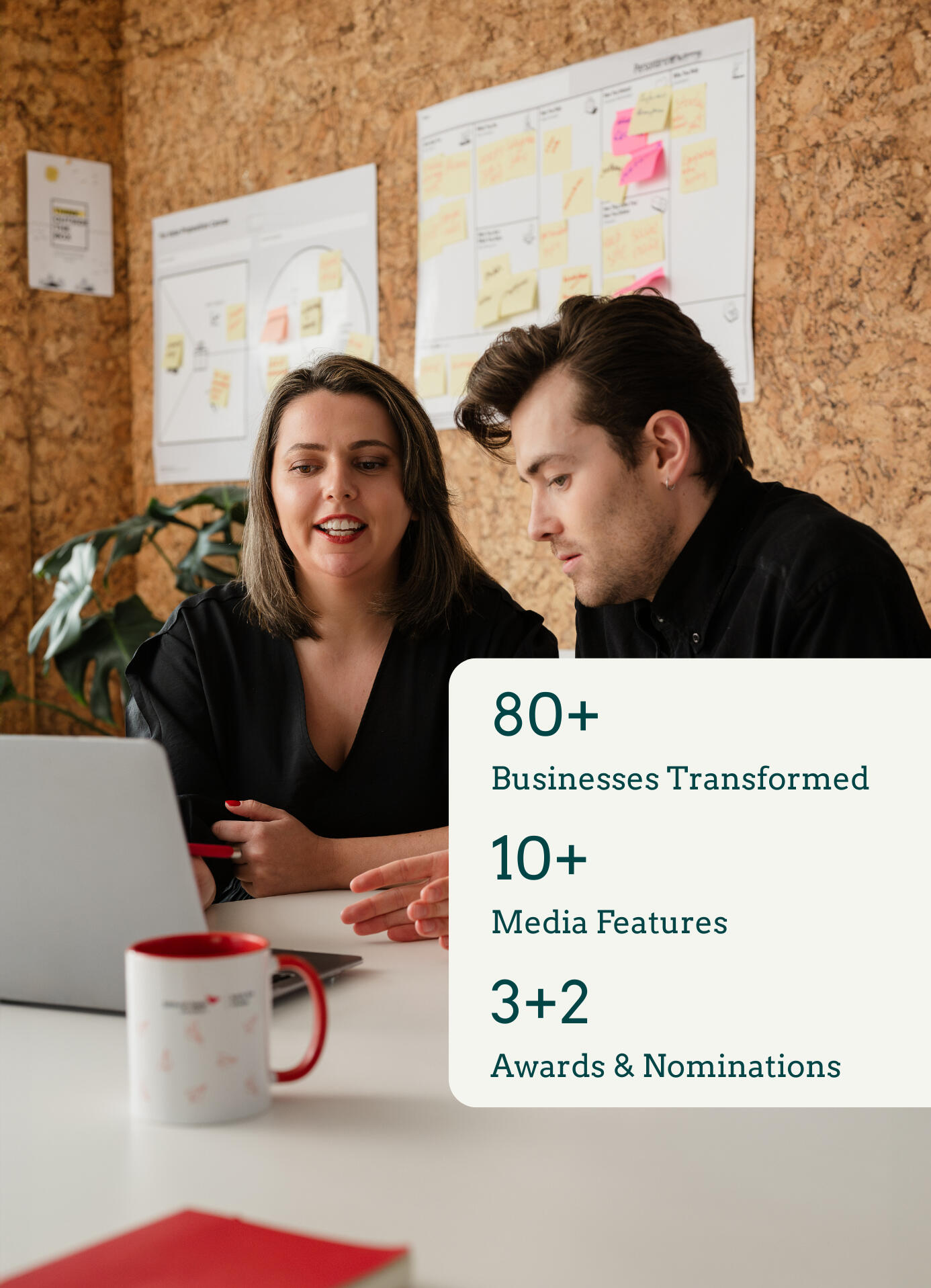 Kitti Majoran is working with a client, sitting at a desk in an office and looking at a laptop. There is also a graphic in the bottom right corner of the photo. On the graphic, the following is written: 70+ businesses transformed, 10+ media features, 3+2 a