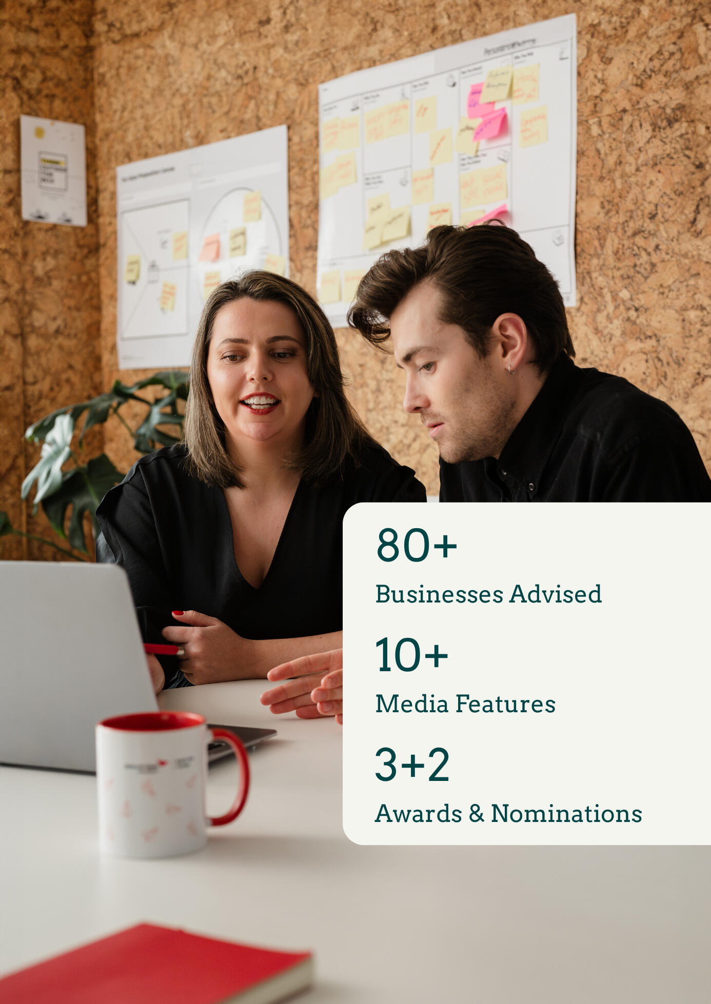 Kitti Majoran is working with a client, sitting at a desk in an office and looking at a laptop. There is also a graphic in the bottom right corner of the photo. On the graphic, the following is written: 80+ businesses transformed, 10+ media features, 3+2 a