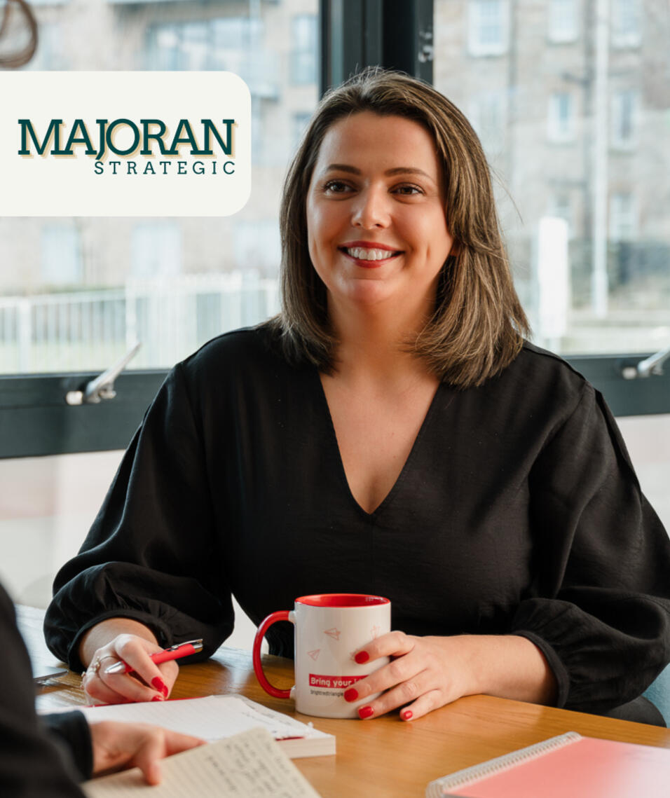 Kitti Majoran is sitting at a desk with a cup of coffee and a pen in her hand, smiling. The KM Strategy logo is also in the top left corner of the photo.